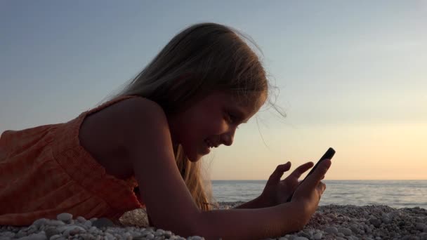 Kid Playing Smartphone, Dziecko na plaży o zachodzie słońca, Młoda Blondynka Korzystanie Tablet na brzegu o zachodzie słońca w wakacje — Wideo stockowe