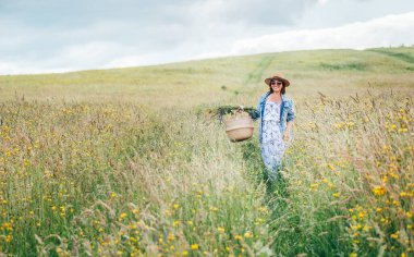 İçtenlikle gülümseyen kot ceket ve açık yaz elbisesi giymiş genç bir kadın sepeti ve kır çiçekleri buketiyle yüksek yeşil çim çayırında yürüyor. İnsan ve doğa kavramı görüntüsü.
