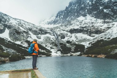 Slovakya 'nın High Tatras kentindeki Velicka vadisinde dağ yürüyüşü yaparken, parlak turuncu ceketli kadın sırt çantasıyla Velicke pleso (dağ gölü) manzarasının tadını çıkarıyor. Doğa konseptinde aktif insanlar.