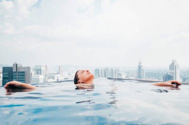 Genç bir bayan şehir merkezindeki gökdelenleri ve kuleleri ile şehir manzaralı sonsuz yüzme havuzunda dinleniyor. Lüks tatil konsepti resmi.