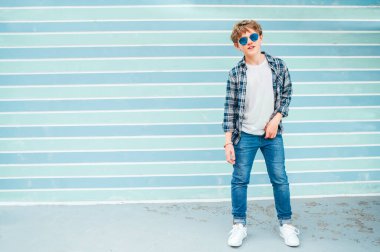 Beyaz sarı saçlı, 12 yaşında, tişörtlü, mavi gözlüklü, turkuaz mavi arka planda poz veren genç bir çocuğun moda portresi. Gençlerin moda konsepti.