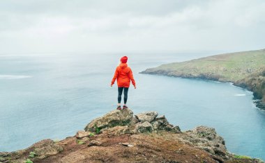 Portekiz 'in Madeira adasının en doğusundaki Ponta de Sao Loureno yarımadasındaki Atlantik Okyanusu manzarasının tadını çıkaran turuncu renkli spor kapüşonlu ve kırmızı şapkalı bir kadın.