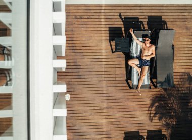 Güverte sandalyesinde güneşlenirken, yüzme şortu, beyzbol şapkası ve güneş gözlüğü takan bir adamın en iyi görüntüsü. Dikkatsiz adam tatil konsepti resmi.