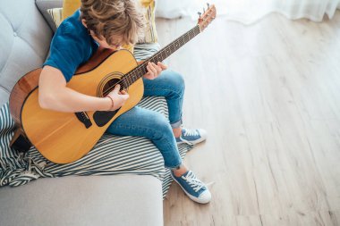 Akustik gitar çalan genç bir çocuk. Günlük kot pantolon, gömlek ve yeni spor ayakkabılar giymiş. Oturma odasındaki rahat koltukta oturuyor. Müzik eğitimi üst açı görünüm konsepti.