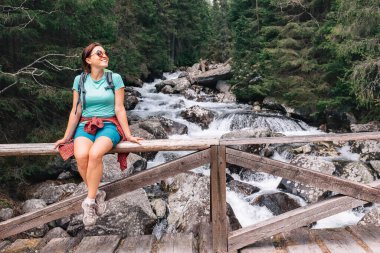 Genç bayan sırt çantalı, soğuk su köprüsünde dinleniyor. Köprü çitlerinde oturan kadın gülümsüyor ve rahatlatıcı anın tadını çıkarıyor. Aktif insanlar doğa konseptinde boş zaman harcıyorlar.