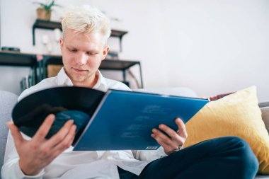 Oturma odasındaki rahat koltukta oturan genç bir adamın portresi. Kolundan 12 inçlik vinil LP çıkarıyor. Eski model analog ses değeri konsept resmi.