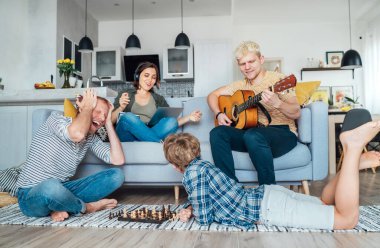 Aile oturma odasında birlikte. Baba oğluyla satranç oynuyor, anne bilgisayarda öğreniyor, büyük oğul akustik gitar çalıyor ve şarkı söylüyor. Karantina zamanı ve aile kavramı.