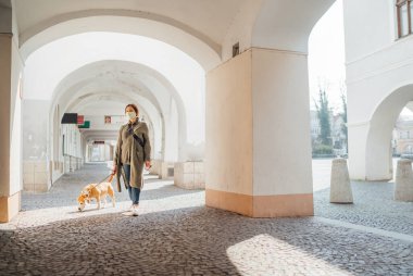 Koronavirüs olarak yüz maskesi takan genç bir kadın terk edilmiş eski Avrupa meydanları ve caddelerinde av köpeğiyle yürümeyi önlüyor. Genel COVID-19 salgın konsept görüntüsü.