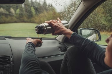 Female pouring the hot tea in tourist thermos mug. She sitting on co-driver seat inside modern car, enjoying the moody rainy day weather looking through windscreen drops. Auto journeys concept image.  clipart