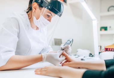 COVID-19 tecrit konseptinde küçük bir işletme var. Şeffaf Güvenli Yüz Kalkanında profesyonel manikür ustası Elektrikli Çivi Kaplama Aracı kullanarak manikür prosedürü. 