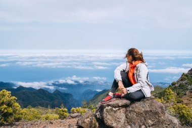 Genç beyaz kadın sportif giysiler giymiş kayanın üzerinde dinleniyor ve Portekiz 'in Madeira Adası' nın en yüksek zirvesi olan Pico Ruivo Dağı 'ndan bulutların manzarasının keyfini çıkarıyor. Etkin tatil kavramı