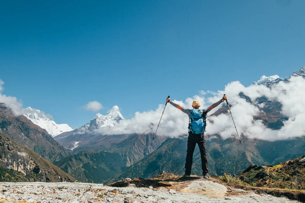 Active vacations concept image. Young hiker backpacker man rising arms with trekking poles enjoying the Thamserku 6608m mountain during high altitude Acclimatization walk. Everest Base Camp route,Nepal.