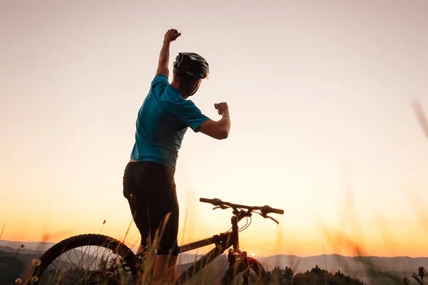 骑自行车的人在山顶与日落相遇 在另一个成功的一天结束时 他举起一只手打招呼 积极运动的人的概念形象 — 图库照片