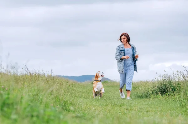 快乐地微笑着 带着飘扬的头发和她的小猎犬跑来跑去 看着眼睛的女慢跑者 与宠物在草原小径上漫步 健康活泼的人的生活方式理念形象 — 图库照片