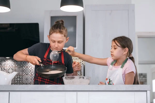 兄と妹は家庭のキッチンで手作りのパンケーキを作るエプロンを着ていました 女の子は熱い鍋に液体生地を入れて キッズホーム料理のコンセプトイメージ — ストック写真