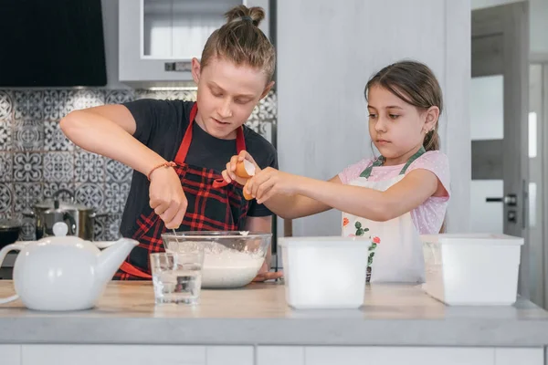 子供家庭料理のコンセプトイメージ 姉と弟は家庭のキッチンで手作りのパンケーキを作るエプロンを着ていました 液体生地と女の子を混合少年は ボウルに卵を破る — ストック写真