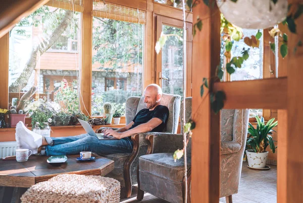 男は快適なアームチェアに座って黒のTシャツとジーンズを着て 現代的なスリムなラップトップを使用し 家のサンルームリビングルームでお茶を飲む 距離またはフリーランスまたはライターの作業コンセプトイメージ — ストック写真