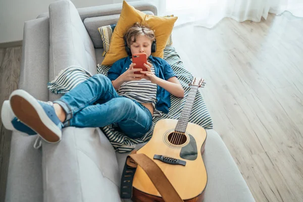 十几岁的男孩躺在舒适的沙发上 穿着休闲裤和新运动鞋 一边听音乐 一边用无线耳机和智能手机聊天 使用电子设备概念 — 图库照片
