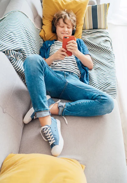 Preteen boy lying at home on cozy sofa dressed casual jeans and new sneakers listening to music and chatting using wireless headphones connected with smartphone. Child use electronic devices concept