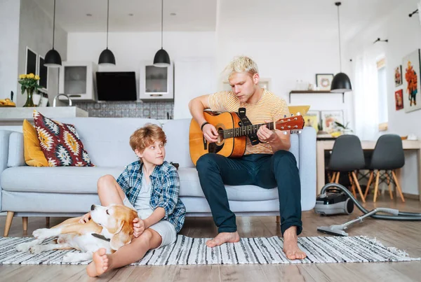 隔離された家族の時間 一緒にリビングルームで小さな兄 ティーンはアコースティックギターを演奏し 床の上で彼らのビーグル犬と遊んで笑顔の小さな男の子 家庭や家族の価値観にとどまる — ストック写真