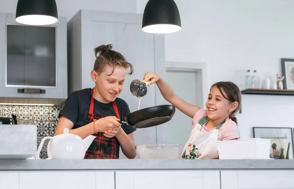 子供家庭料理のコンセプトイメージ 姉と弟は家庭のキッチンで手作りのパンケーキを作るエプロンを着ていました 女の子ポルノの液体生地上にザ ホットパン — ストック写真