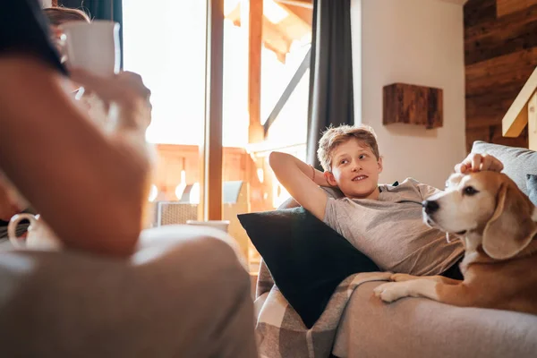 Cozy family tea time. Father and son at the home living room. Boy lying on comfortable sofa and  stroking their beagle dog and smiling. Peaceful family moments concept image.