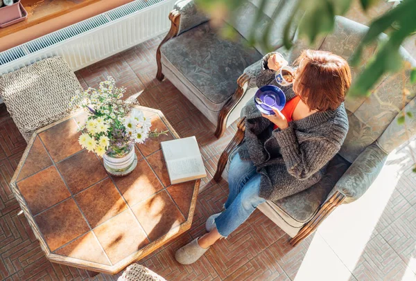 快適なアームチェアに座っている居心地の良い家庭の服を着た女性のトップビューと彼女は本を読んでいる間 家のサンルームリビングルームで朝のコーヒーを飲みます — ストック写真