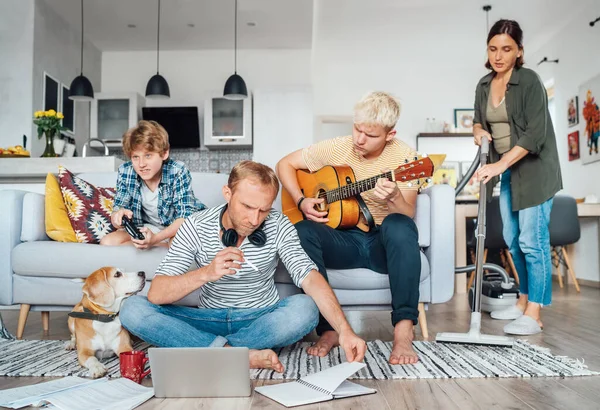 Family at living room together on Quarantine time.Father distance working using laptop, mother cleaning the room, older son playing acoustic guitar and singing a songs,little son playing video concept