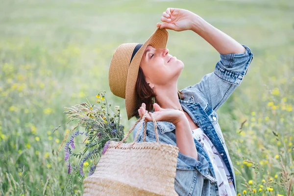 一个年轻女人的画像闭上了眼睛 欣赏着阳光 她穿着牛仔裤夹克 草帽和浅色夏装 她在草地上摘了一朵野花 把花束放在篮子里 — 图库照片