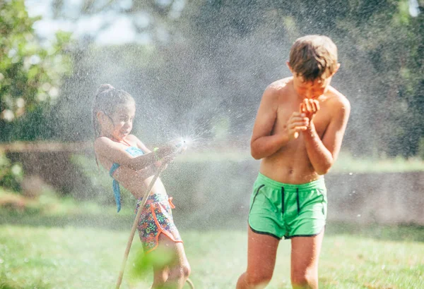 男の子と女の子は庭で遊んで ホースから互いに水を注ぎ 雨を降らせます 幸せな子供時代のコンセプトイメージ — ストック写真
