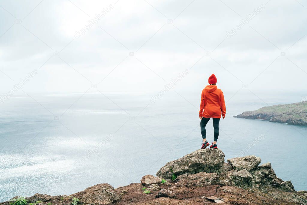 Trail runner female dressed orange sporty hoodie and red cap resting on the cliff and enjoying Atlantic ocean view on San Lourenco peninsula the easternmost point of Madeira island, Portugal