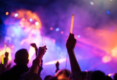 Genç kız hayranı, gece konserinde en sevilen popüler grubu destekleyen renkli çubuklarla iki elini kaldırdı. Işık ve lazer gösterisiyle renkli kalabalık arka plan eğlencesi. 