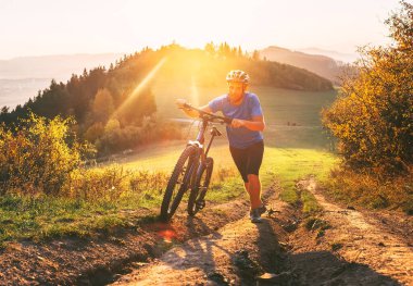 Tepeye dağ bisikletini iten gülümseyen genç bir adam. Bisikletle aktif macera yolculuğu.