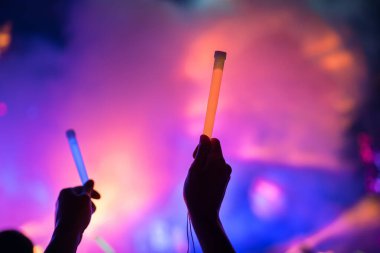 Genç kız hayranı, gece konserinde en sevilen popüler grubu destekleyen renkli çubuklarla iki elini kaldırdı. Işık ve lazer gösterisiyle renkli kalabalık arka plan eğlencesi. 