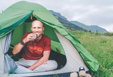 Yeşil çadırda içtenlikle gülümseyen adam sisli bir sabah manzarası olan termostan kahve içiyor. Aktif insanlar çadırların konsept imajıyla seyahat etmekten hoşlanıyor.