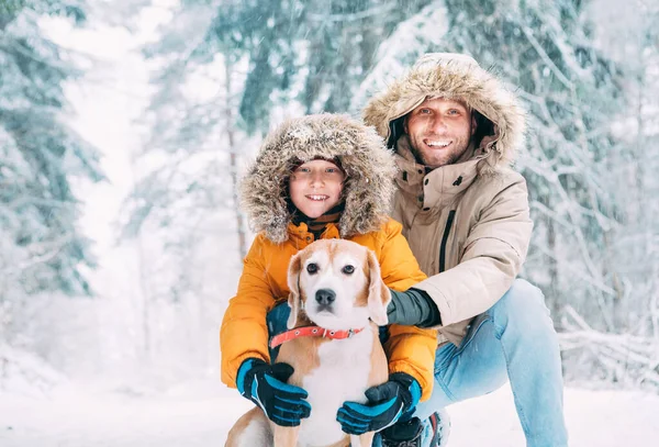 父亲和儿子穿着保暖帽衫 带着他们的小猎犬在雪地的森林里散步 笑容满面 宠物在家庭和冬季服装的概念形象 — 图库照片