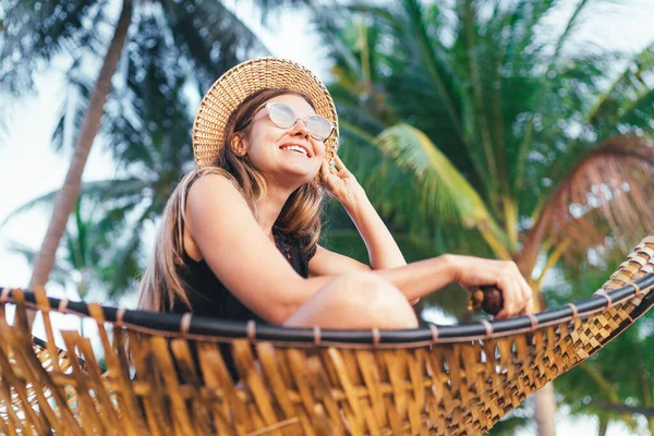 戴着太阳镜戴着草帽 笑容满面的年轻女子坐在带有棕榈树背景的吊床上 — 图库照片