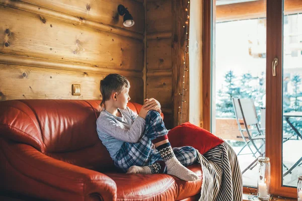 Boy sitting on cozy couch at living room and watching in wide window in cozy home atmosphere. Peaceful moments of cozy home concept image.