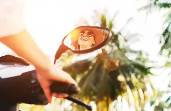 Smiling woman riding motorbike mirrored in rearview mirror with shining sunrays on the background.Cheerful people during vacation time concept image