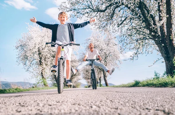 広い足を広げ 桜の木の下の田舎道で自転車に乗るときに叫んで楽しんでいる父と息子 健康的なスポーティライフスタイルのコンセプトイメージ — ストック写真