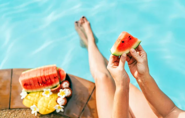 女性は小さなスイカの作品を手に取り プールの端に座って新鮮な果物を楽しむ — ストック写真