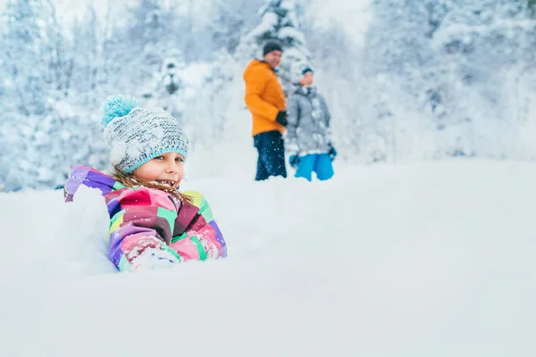 在冬季雪地的森林里和家人一起散步时 快乐地微笑着的小女孩坐在厚厚的雪地里 — 图库照片