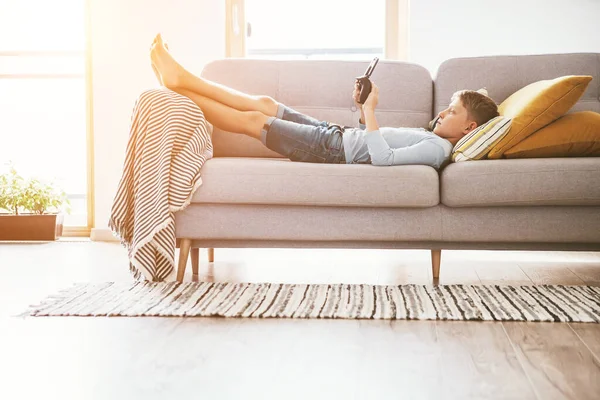 男の子は電子機器で遊ぶ ゲームパッド自宅のリビングルームの居心地の良いソファに横たわってスマートフォンに接続 — ストック写真