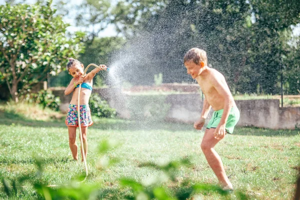 庭で遊んでいる2人の子供は ホースからお互いを注ぎ 雨を降らせます 幸せな子供時代のコンセプトイメージ — ストック写真