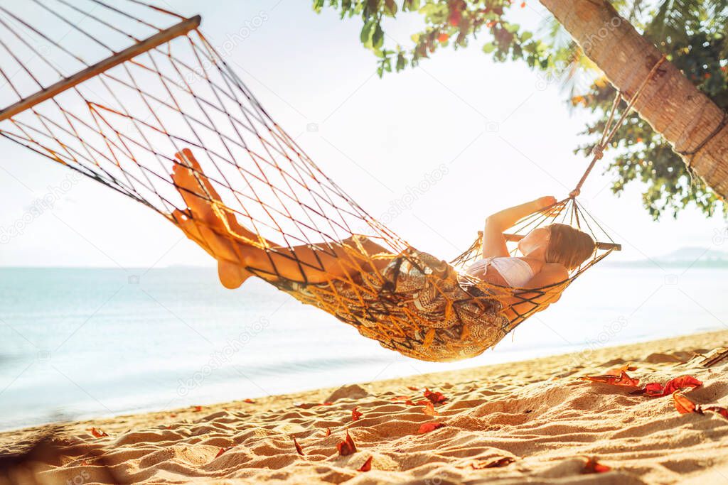 Young blonde longhaired woman relaxing in hammock hinged between palm trees on the sand beach 