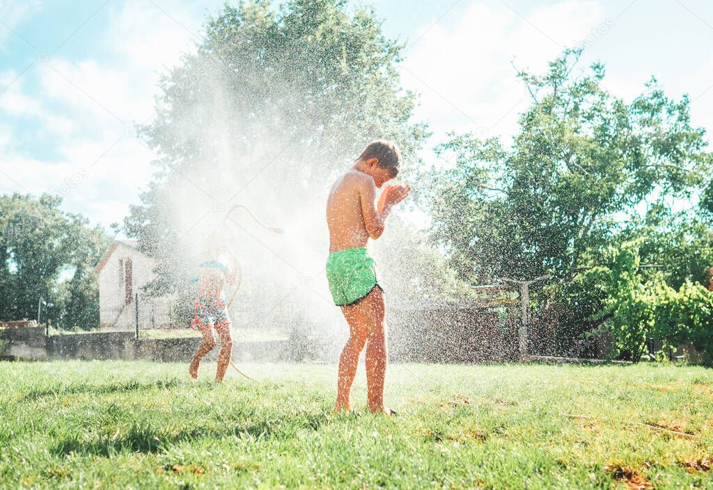 Hot summer days activity: happy little girl jumps under water, when brother pours her from garden hose