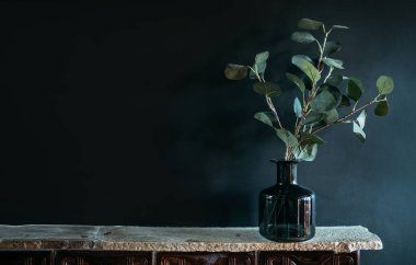 Yeşil ağaç dalı, yan pencere ışığıyla aydınlatılan siyah duvar zeminindeki doğal taş şömine rafındaki siyah cam vazoya kondu. Konforlu ev dekorasyonu unsurları konsept resmi.