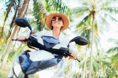 Hasır şapkalı ve güneş gözlüklü mutlu bir kadın palmiye ağacının altında motosiklet sürüyor.