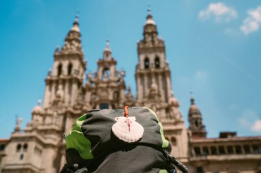 Pilgrim's backpack with famous pilgrims' mascot and sign seashell with Cross of Saint James at  on the Obradeiro square (plaza) - the main square in Santiago de Compostela with Catedral de Santiago. clipart