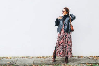 Gülümseyen kadın, boho tarzı renkli uzun bir elbise, siyah deri ceketli, kahverengi deri çantalı, beyaz arka planda poz veren..